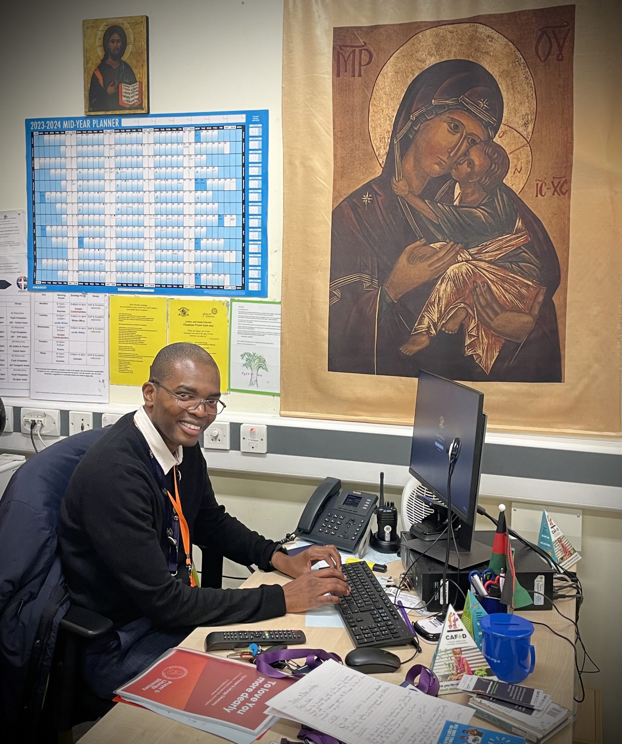 Deacon Davie sits at his desk in front of his computer, smiling at the camera. In the background, the is a large poster depicting Our Lady holding the infant Jesus