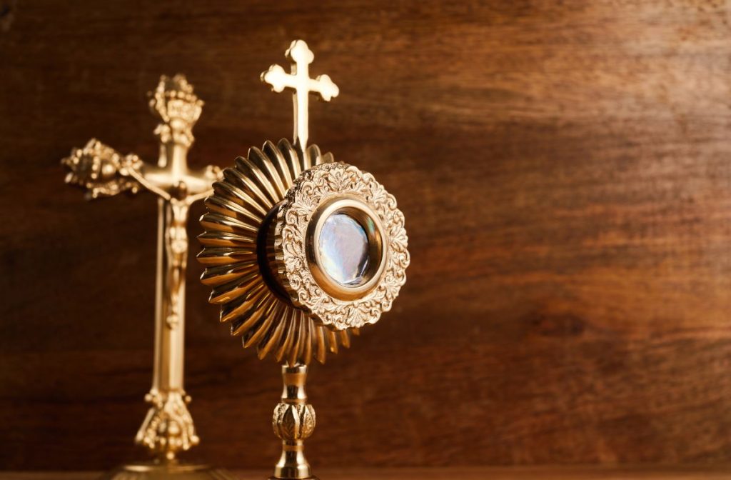A golden monstrance with Eucharist in the centre and a golden cross on top. Behind is a large golden crucifix. 