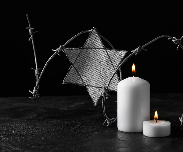 Two lit white candles flicker against a dark background along with a grey Star of David surrounded with barbed wire