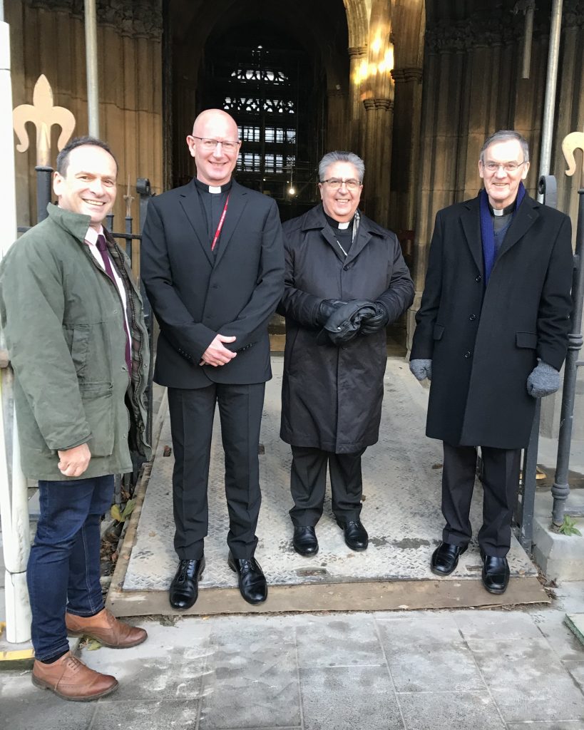 Chris Cotton, from Purcell Architects, Canon Michael Jones, Archbishop Buendia, and Bishop John stand outside the door to the Cathedral. A stained glass window can be seen in the background.