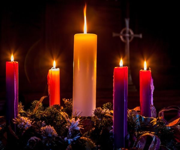 Three purple candles, one pink, and one white, are lit and arranged inside an Advent wreath, set against a dark background